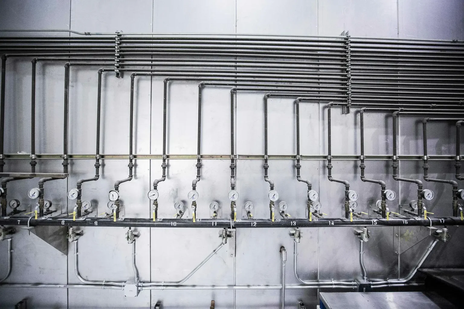 System av industriella rör med numrerade tryckmätare och ventiler på en metallvägg